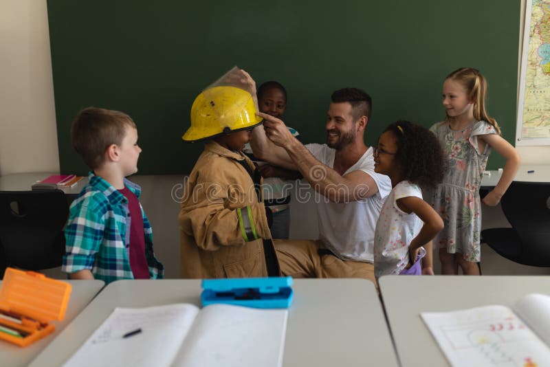 Front view of male Caucasian firefighter helps to wearing fire uniform to schoolkid with all the schoolkids around in classroom of elementary school. Front view of male Caucasian firefighter helps to wearing fire uniform to schoolkid with all the schoolkids around in classroom of elementary school