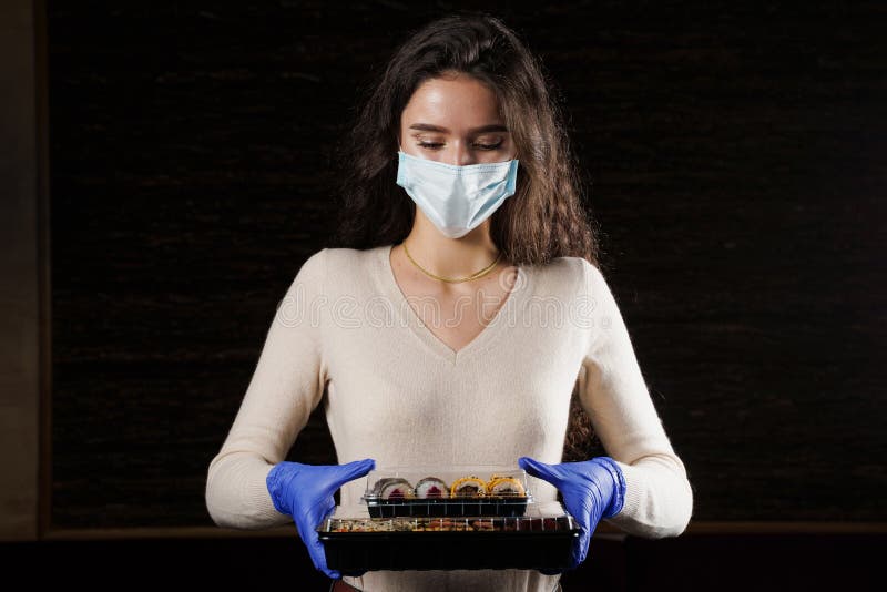 Girl with sushi. Food courier in medical mask. Delivery at quarantine coronavirus covid-19. Young woman holding 2 sushi boxes in hands using gloves. Girl with sushi. Food courier in medical mask. Delivery at quarantine coronavirus covid-19. Young woman holding 2 sushi boxes in hands using gloves