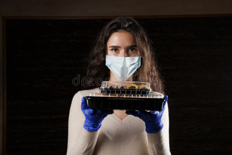 Girl with sushi. Food courier in medical mask. Delivery at quarantine coronavirus covid-19. Young woman holding 2 sushi boxes in hands using gloves. Girl with sushi. Food courier in medical mask. Delivery at quarantine coronavirus covid-19. Young woman holding 2 sushi boxes in hands using gloves