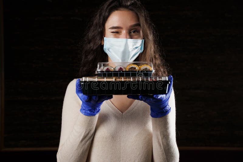 Girl with sushi. Food courier in medical mask. Delivery at quarantine coronavirus covid-19. Young woman holding 2 sushi boxes in hands using gloves. Girl with sushi. Food courier in medical mask. Delivery at quarantine coronavirus covid-19. Young woman holding 2 sushi boxes in hands using gloves