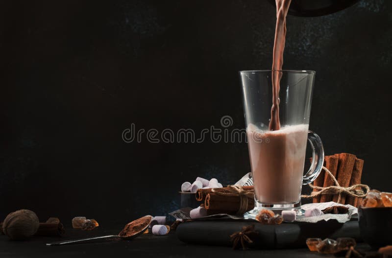 Pouring winter hot chocolate cocoa drink into tall glass with ispices on black table. Dark blue background. Copy space. Pouring winter hot chocolate cocoa drink into tall glass with ispices on black table. Dark blue background. Copy space