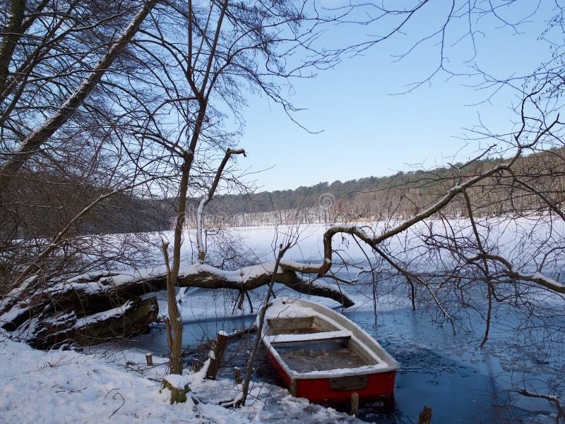 Small wooden rowboat in a frozen lake in winter with leafless deciduous tree branches and a snowy mountainous landscape. Small wooden rowboat in a frozen lake in winter with leafless deciduous tree branches and a snowy mountainous landscape
