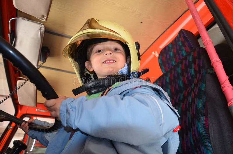 Little boy acting like a fireman. Boy sitting in a real fireman car. Happy adorable child boy with fireman hat sitting in red fire truck looking out. Dreaming of future profession. Fire safety, life protection lessons. Little boy acting like a fireman. Boy sitting in a real fireman car. Happy adorable child boy with fireman hat sitting in red fire truck looking out. Dreaming of future profession. Fire safety, life protection lessons.