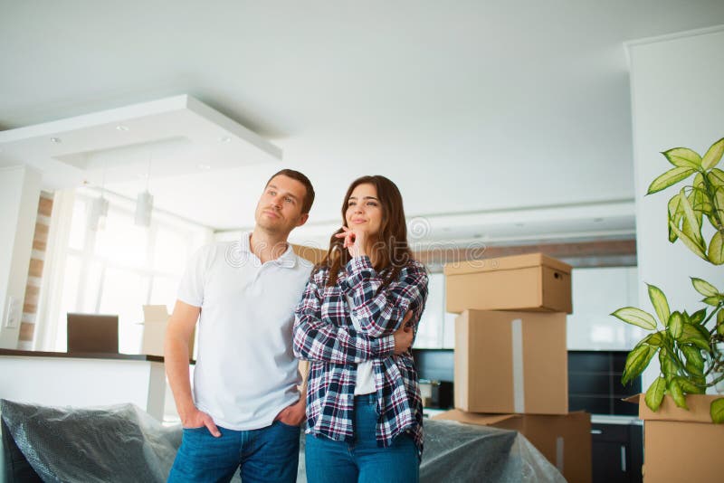 A young married couple in the living room in the house stand near unpacked boxes. hey are happy about new home. Moving, buying a house, apartment concept. A young married couple in the living room in the house stand near unpacked boxes. hey are happy about new home. Moving, buying a house, apartment concept