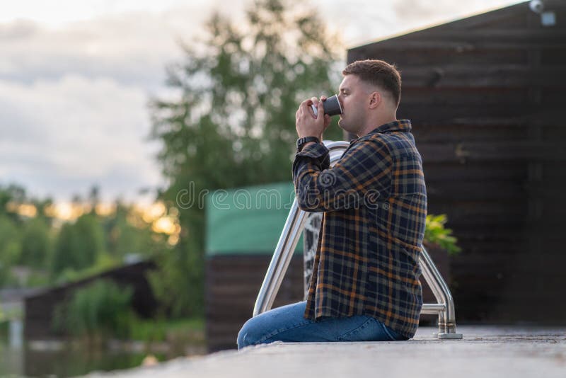 Young man gulping down cup of refreshing takeaway coffee as he relaxes on a wooden jetty at sunset. Young man gulping down cup of refreshing takeaway coffee as he relaxes on a wooden jetty at sunset