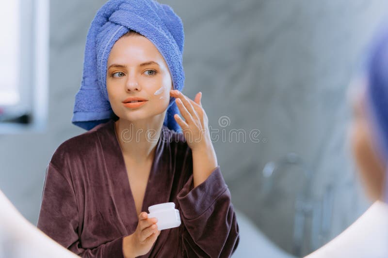 portrait of young woman looking in mirror, applying moisturizing cream lotion on cheeks, Morning skin care at home. Smiling woman taking care of herself after showering in the bathroom. portrait of young woman looking in mirror, applying moisturizing cream lotion on cheeks, Morning skin care at home. Smiling woman taking care of herself after showering in the bathroom.