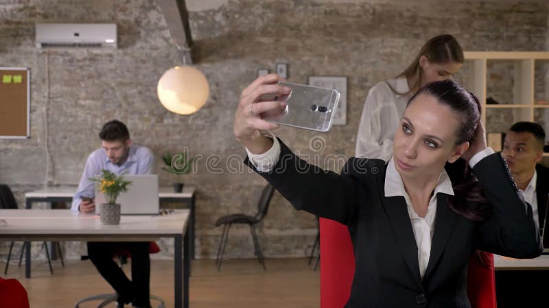 年轻女实业家在智能手机做selfie在办公室，她的同事是与技术的网络，运作