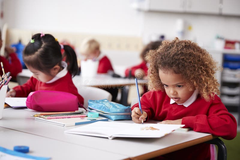 年轻女小学生佩带的校服坐直在一张幼儿学校教室图画的一张书桌的,关闭 免版税图库摄影 年轻女小学生佩带的校服坐直在一张幼儿学校教室图画的一张书桌的,关闭 免版税图库摄影