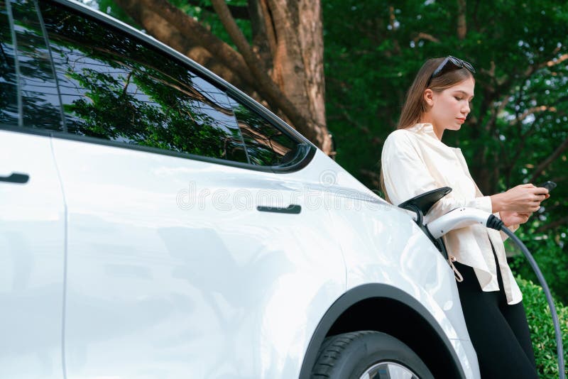 Young woman use smartphone to pay for electricity at public EV car charging station green city park. Modern environmental and sustainable urban lifestyle with EV vehicle. Expedient. Young woman use smartphone to pay for electricity at public EV car charging station green city park. Modern environmental and sustainable urban lifestyle with EV vehicle. Expedient