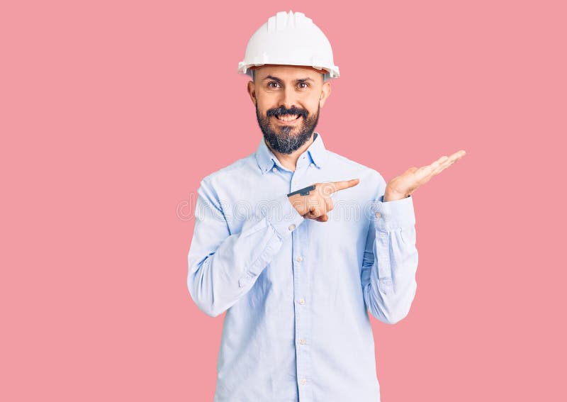 年轻帅哥戴着建筑师精装帽，一边用手和手指指着相机，一边惊叹微笑 库存图片