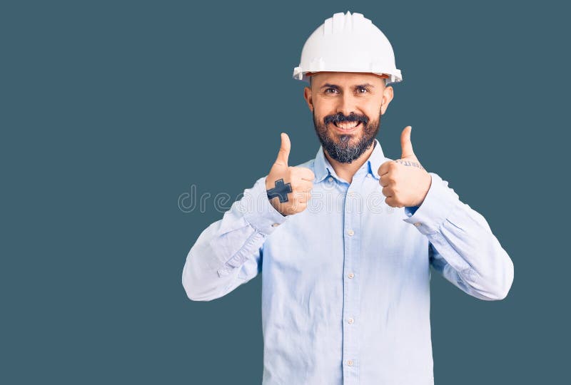 年轻帅哥穿着建筑师精装成功标志用手拇指微笑开心地做正面手势 免版税库存图片