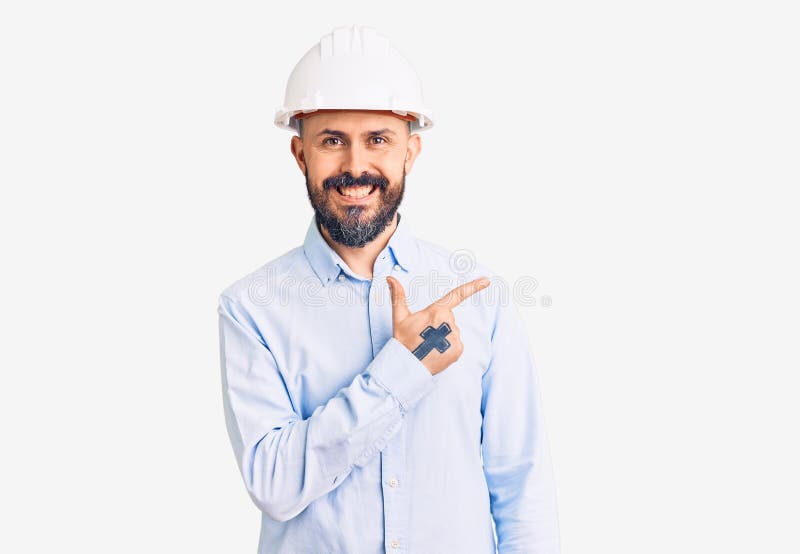 年轻帅气的建筑师戴着精装帽，脸上带着笑脸，手指朝上，高兴地指着身边 免版税库存图片