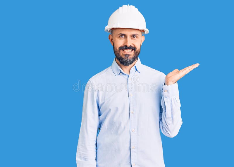 年轻帅气的男子，戴着建筑师的硬帽，微笑着欢快地呈现，并用手掌指着镜头 库存照片