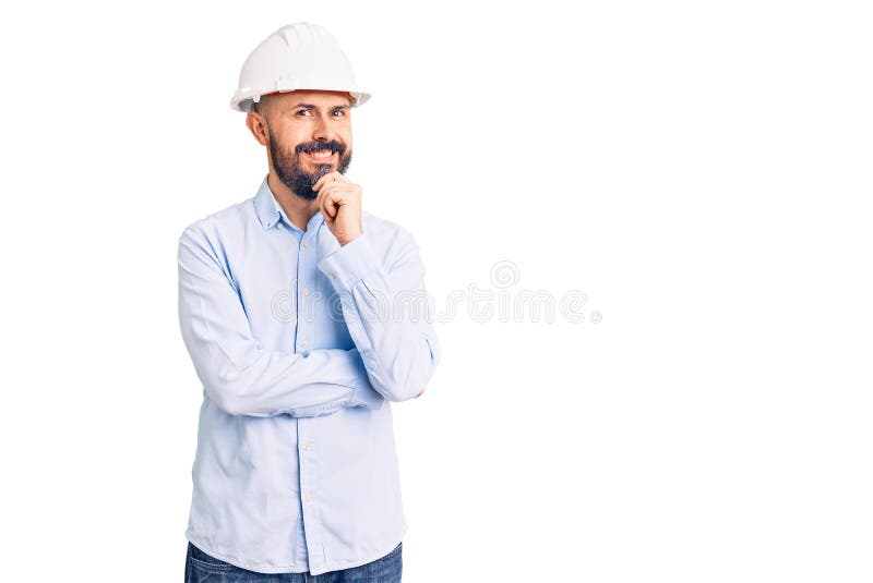 年轻帅气的男子，戴着建筑师的硬帽，面带微笑，双臂交叉，手举在下巴上，面带自信 库存照片