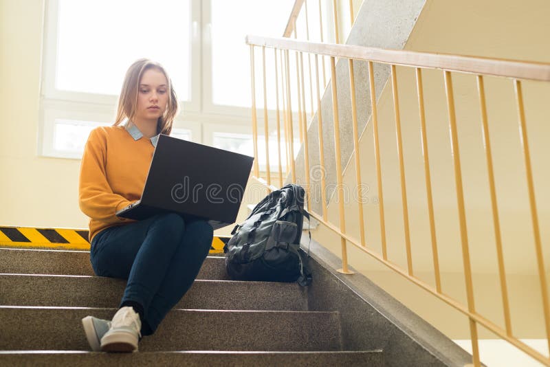 Young depressed lonely female college student sitting on stairs at her school, writing on her laptop. Education, Bullying, Depression concept. Young depressed lonely female college student sitting on stairs at her school, writing on her laptop. Education, Bullying, Depression concept.