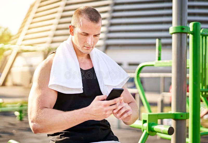 年轻的20多岁肌肉运动员在脖子上用毛巾进行锻炼，在户外用智能手机进行锻炼之间休息 免版税库存照片