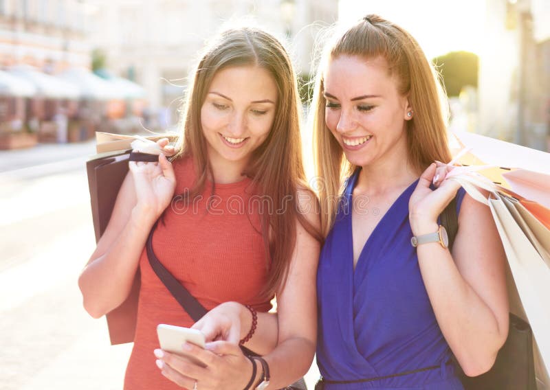 年轻的快乐千禧朋友带着购物袋走在街上查看社交媒体平台更新了两名学生 免版税库存照片