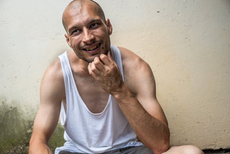 年轻皮包骨头的厌食秃头正面和愉快的微笑的无家可归的人坐都市街道在城市或镇在白色墙壁附近 库存照片 年轻皮包骨头的厌食秃头正面和愉快的微笑的无家可归的人坐都市街道在城市或镇在白色墙壁附近 库存照片