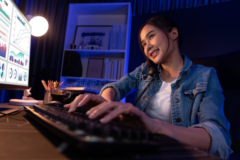 Young beautiful Asian creative woman calling on phone to customer or coworker informing strategy marketing creator plan while searching market analysis on pc at neon modern office at night. Stratagem. Young beautiful Asian creative woman calling on phone to customer or coworker informing strategy marketing creator plan while searching market analysis on pc at neon modern office at night. Stratagem.