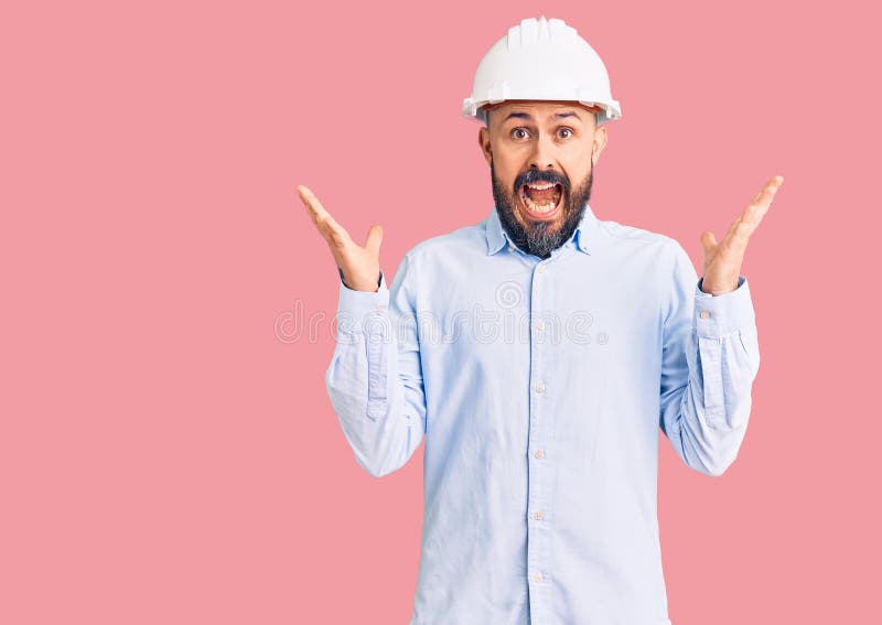 年轻英俊男子戴着建筑师精装帽，为疯狂而欢呼，并惊讶于成功，他举起双臂，睁开双眼尖叫 免版税库存照片