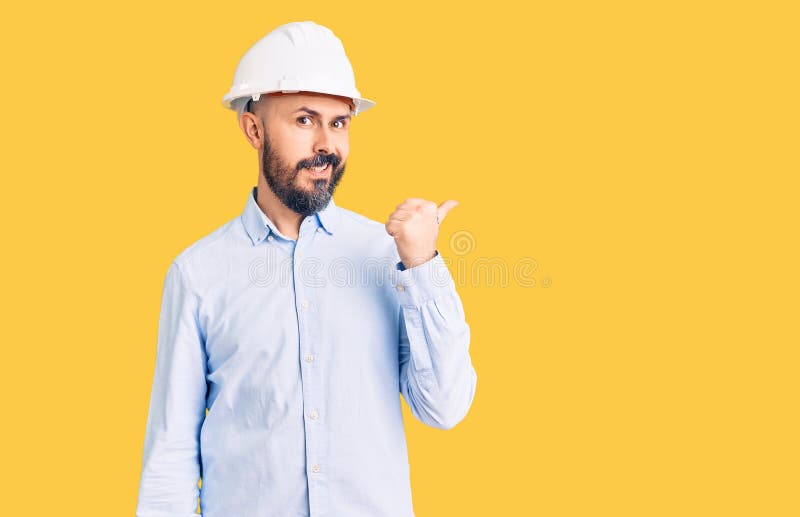 年轻英俊的男子戴着建筑师的硬帽，面带快乐的脸孔微笑，用大拇指朝侧指 免版税图库摄影