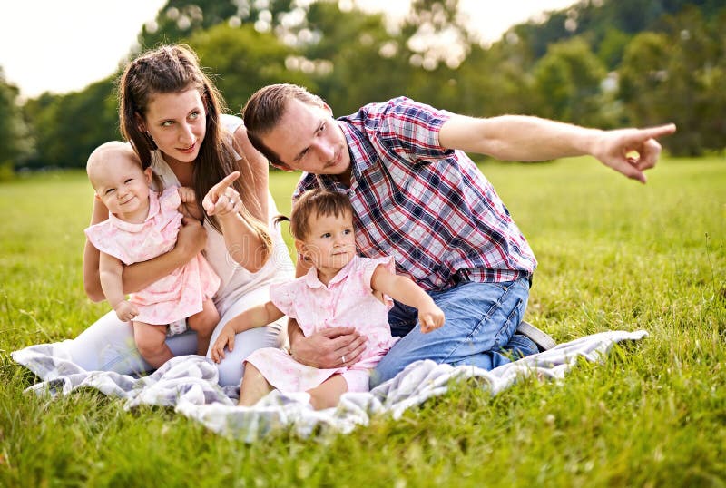 幸福的家庭，年轻的千禧一代父母和两个女婴，坐在野餐毯上，指着一些东西 免版税图库摄影