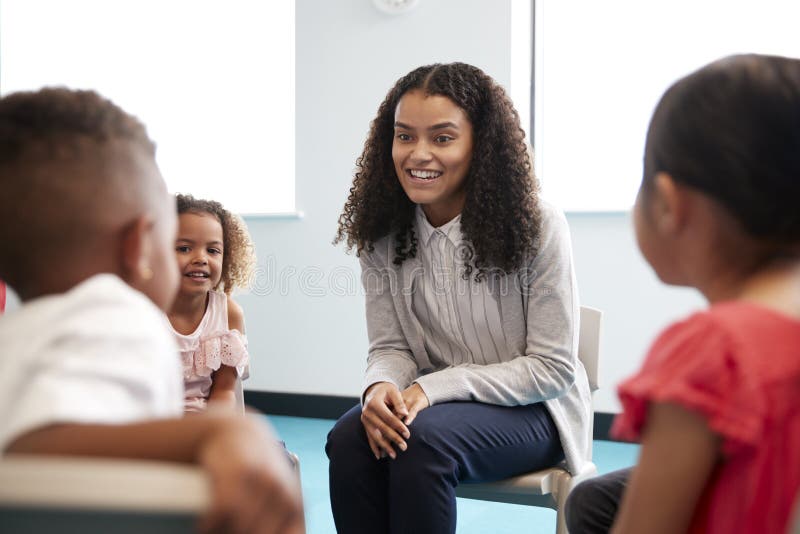 幼儿学校孩子坐在一个圈子的椅子在教室谈话与他们微笑的女老师,关闭,有选择性的f 免版税库存图片 幼儿学校孩子坐在一个圈子的椅子在教室谈话与他们微笑的女老师,关闭,有选择性的f 免版税库存图片