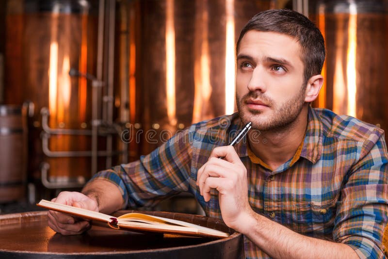 Thoughtful young man in casual shirt holding note pad and looking away while leaning at the wooden barrel with metal containers in the background. Thoughtful young man in casual shirt holding note pad and looking away while leaning at the wooden barrel with metal containers in the background