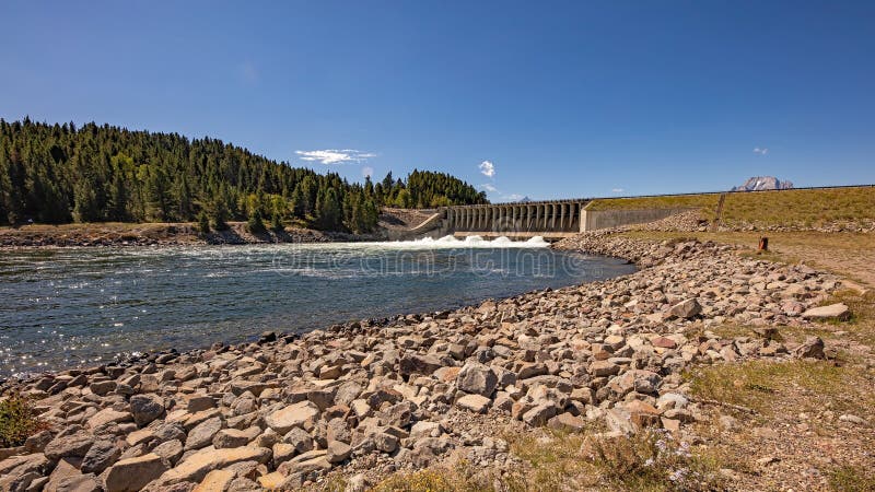 Water spills from the dam on the Snake River which backs up water to form Jackson Lake. Rushing water continues to flow toward Idaho from the mountains of Yellowstone National Park in Wyoming. The peaks of Mount Moran and Grand Teton can just be seen over the top edge of the dam. Large rocks have been placed to line the shore to prevent erosion. Some haze from smoke from Canadian wildfires can be seen lining the horizon. Under a clear blue sky in early September. Water spills from the dam on the Snake River which backs up water to form Jackson Lake. Rushing water continues to flow toward Idaho from the mountains of Yellowstone National Park in Wyoming. The peaks of Mount Moran and Grand Teton can just be seen over the top edge of the dam. Large rocks have been placed to line the shore to prevent erosion. Some haze from smoke from Canadian wildfires can be seen lining the horizon. Under a clear blue sky in early September.