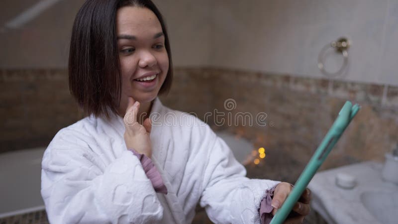 微笑的小女子在家中的浴室里,用平板电脑触摸面部皮肤. 自信的正面肖像 影视素材