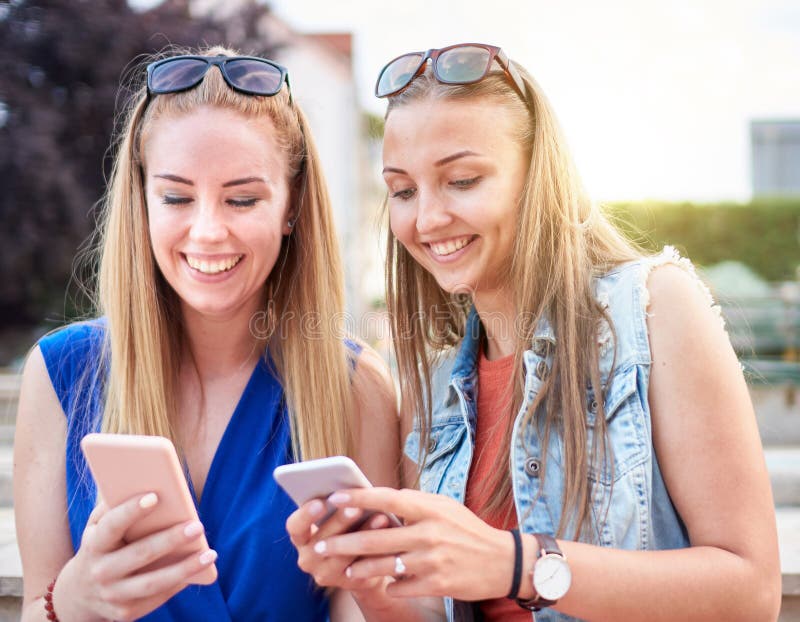 微笑的年轻千禧一代女性朋友在智能手机上查看社交媒体更新 - 两名快乐的女性坐在长椅上，并且 库存照片