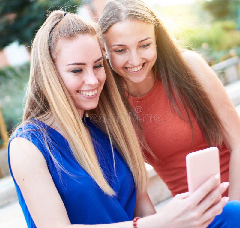 微笑的年轻千禧女朋友在社交媒体上查看最新消息在智能手机上：两个坐在长凳上的幸福女人 库存图片