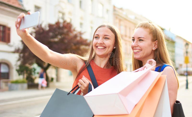 快乐的千禧女朋友自拍玩得开心地和彩纸购物袋一起走在街上 库存图片