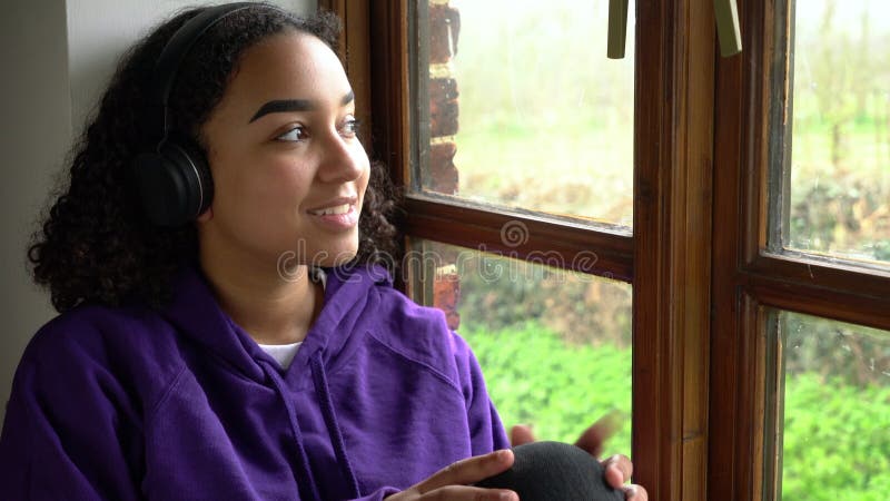 快乐的女青少年双种族裔非洲裔少女，坐在窗边，戴着无线耳机听音乐