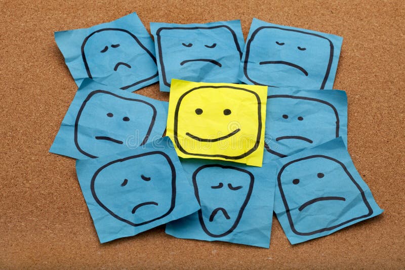 Positive attitude or optimism concept - happy smiley face on yellow sticky note surrounded by sad unhappy blue faces. Positive attitude or optimism concept - happy smiley face on yellow sticky note surrounded by sad unhappy blue faces