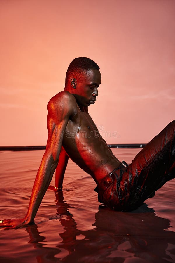 sexy shirtless african american man in wet black pants sitting on water surface in red lights, stock photo. sexy shirtless african american man in wet black pants sitting on water surface in red lights, stock photo