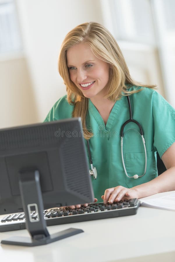 愉快的医生In Scrubs在医院书桌的Using Computer