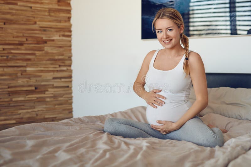 Happy pregnant woman sitting on a bed cradling her baby bump with her hands smiling at the camera. Happy pregnant woman sitting on a bed cradling her baby bump with her hands smiling at the camera