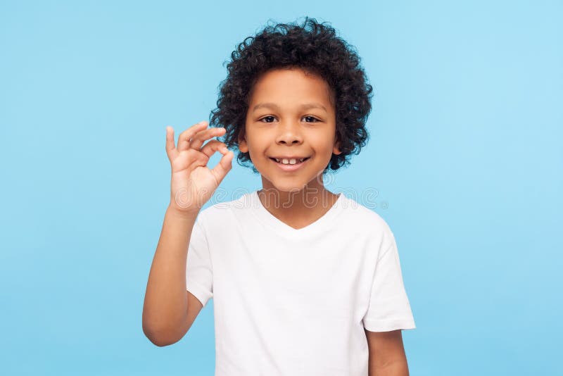 I`m okay! Portrait of adorable joyful little boy with curly hair in white T-shirt showing ok hand gesture and smiling, healthy happy contented with life. indoor studio shot isolated on blue background. I`m okay! Portrait of adorable joyful little boy with curly hair in white T-shirt showing ok hand gesture and smiling, healthy happy contented with life. indoor studio shot isolated on blue background
