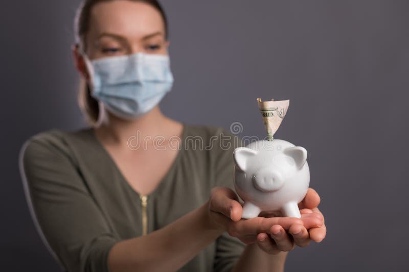 戴冠状病毒口罩的女孩，拥有小猪存钱罐和美元 省钱，经济衰退 有关标题、新闻和 免版税库存照片
