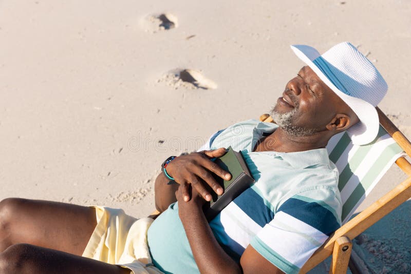 High angle view of african american senior man wearing hat with eyes closed relaxing on deckchair. Beach, summer, book, sunbathing, unaltered, lifestyle, vacation, retirement, enjoyment and nature. High angle view of african american senior man wearing hat with eyes closed relaxing on deckchair. Beach, summer, book, sunbathing, unaltered, lifestyle, vacation, retirement, enjoyment and nature.