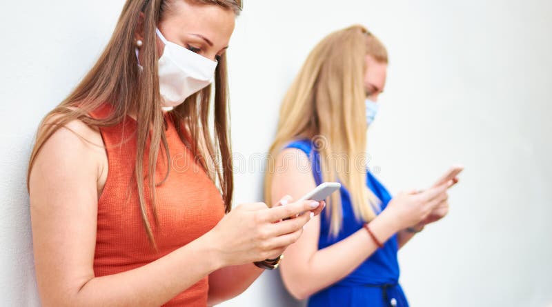 戴着口罩的年轻女朋友使用社交媒体网络和移动智能手机，千禧一代女孩保持距离 库存图片