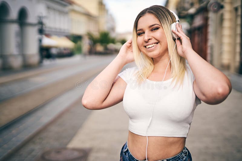 戴着头戴耳机、头像上印着少年头像的城市街道上，一位身着大衣、听音乐的无忧无虑的千禧女性 免版税库存图片