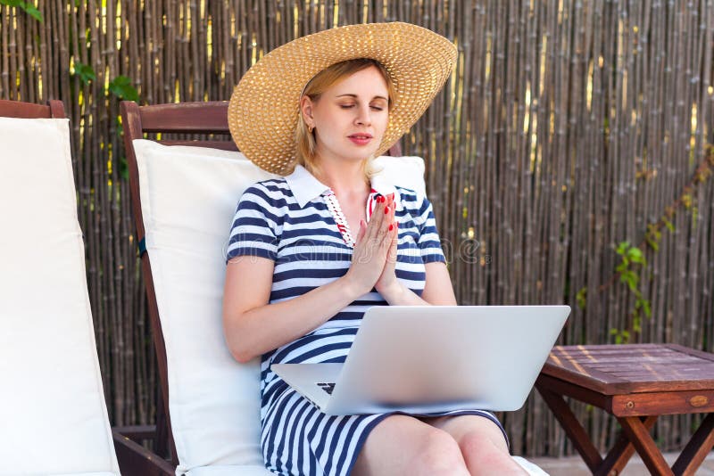 Portrait of beautiful calm young adult freelancer prayer woman in hat and dress is sitting on cozy sunbed with laptop and praying with closed eyes, asking good career. Lifestyle, outdoor, summer. Portrait of beautiful calm young adult freelancer prayer woman in hat and dress is sitting on cozy sunbed with laptop and praying with closed eyes, asking good career. Lifestyle, outdoor, summer