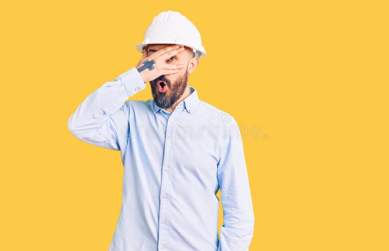 戴着建筑师硬帽的年轻英俊男子，用手指透过手指露出震惊的脸和眼睛 免版税库存照片