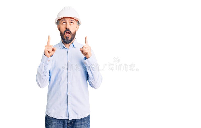 戴着建筑设计精装帽的年轻英俊男子惊讶地看着和指指点点，举起双臂 免版税图库摄影