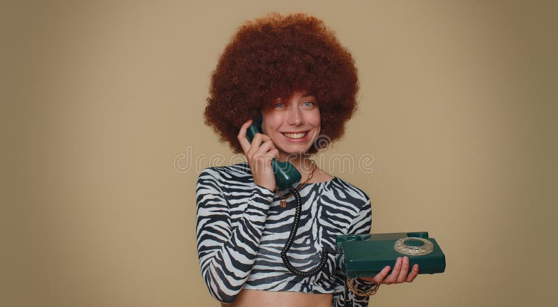 Hey you, call me back. Pretty young woman with brown lush wig talking on wired vintage telephone of 80s, says hey you call me back. Adult stylish female girl isolated on beige studio background indoor. Hey you, call me back. Pretty young woman with brown lush wig talking on wired vintage telephone of 80s, says hey you call me back. Adult stylish female girl isolated on beige studio background indoor