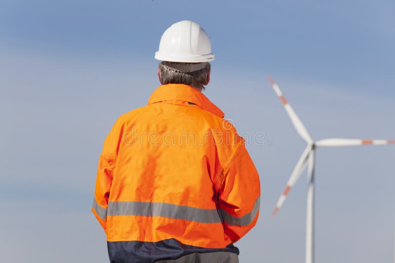Worker or engineer with hard hat and protecive clothing looking at a windmill of a windfarm - focus on the man. Worker or engineer with hard hat and protecive clothing looking at a windmill of a windfarm - focus on the man