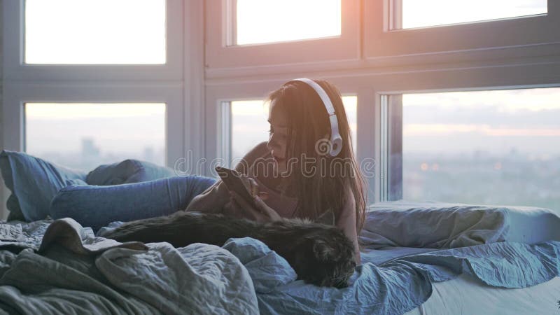 戴耳机的年轻可爱女人，与床上可爱的小猫放松，听窗边的音乐唱歌