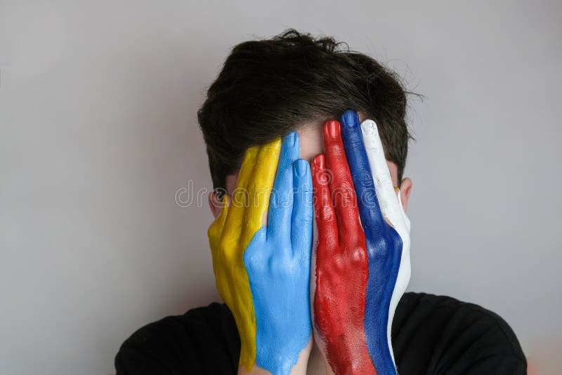 Hand painted in the colors of the flag of Russia and Ukraine. War in Ukraine. Concept of war between Ukraine and Russia. The palms cover the young man&#x27;s face. Hand painted in the colors of the flag of Russia and Ukraine. War in Ukraine. Concept of war between Ukraine and Russia. The palms cover the young man&#x27;s face.